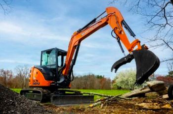 A compact excavator with a hydraulic thumb, working beside a pile of logs