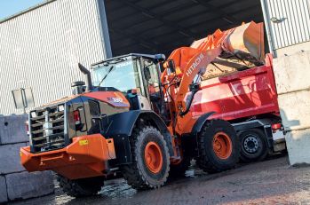A Hitachi/Landcros ZW180 wheel loader dumping a bucket into a red dump truck